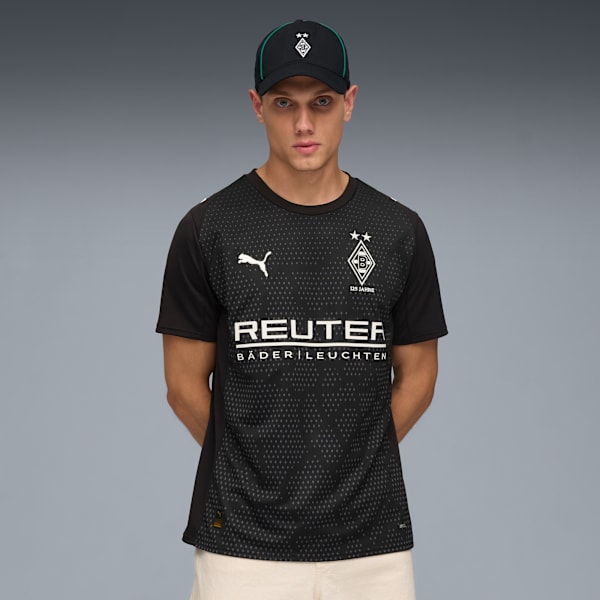 Against a grey backdrop, a model wears a cap and a black patterned soccer jersey.