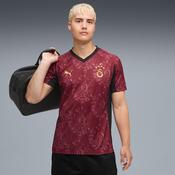 A model carries a black duffel bag, wearing a patterned maroon football shirt against grey.