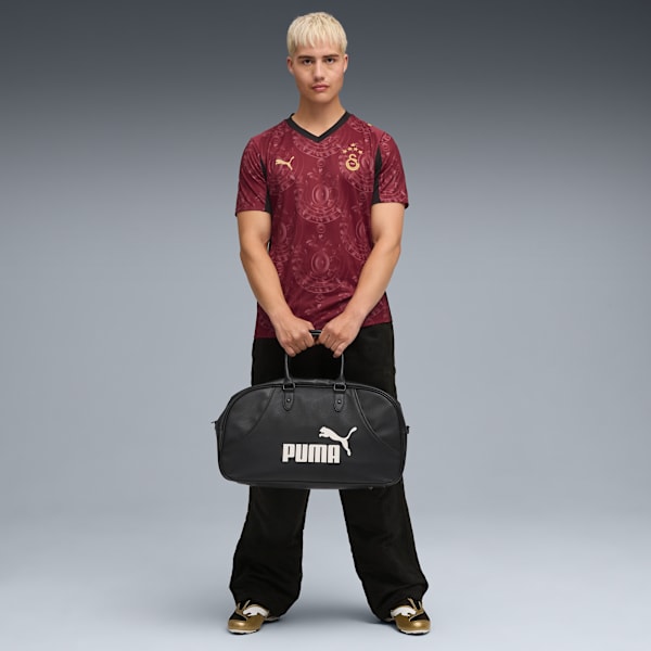 A model poses with a black bag in front of a grey background wearing a patterned maroon t-shirt.