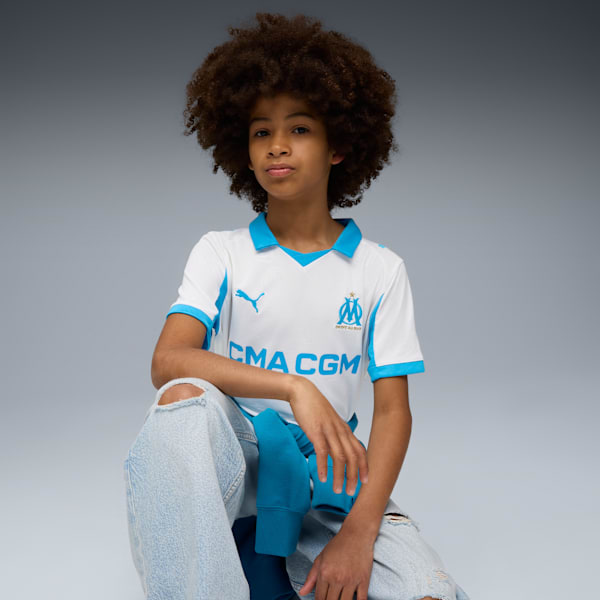 On grey, a model kneels, wearing a white and blue football shirt and ripped denim jeans.