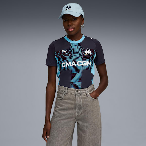 A model poses against a grey backdrop, wearing a cap, football shirt and grey jeans.