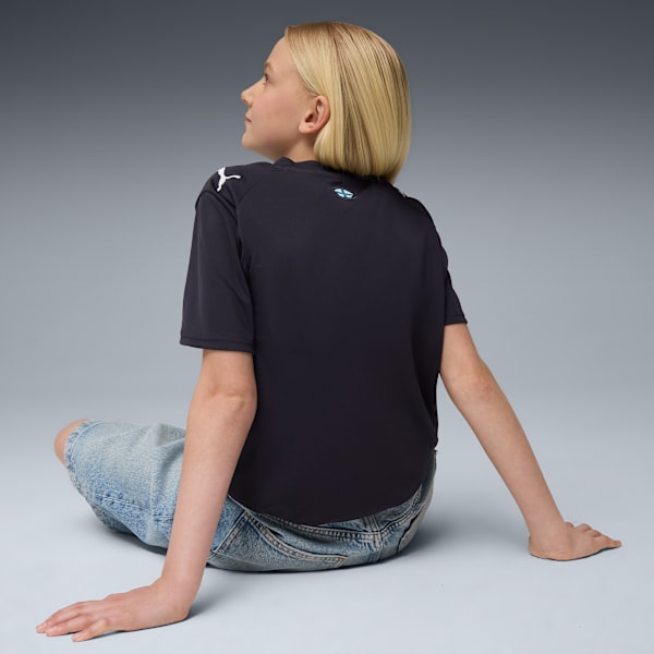 A model with blonde hair sits against grey wearing a dark t-shirt and blue jeans.