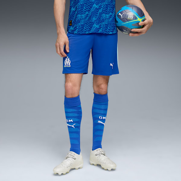 On a grey background, a model holds a ball, wearing blue shorts, socks and sports shoes.