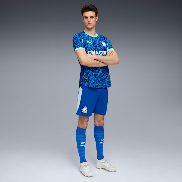 A model stands against grey wearing a blue patterned shirt, shorts, socks and trainers.