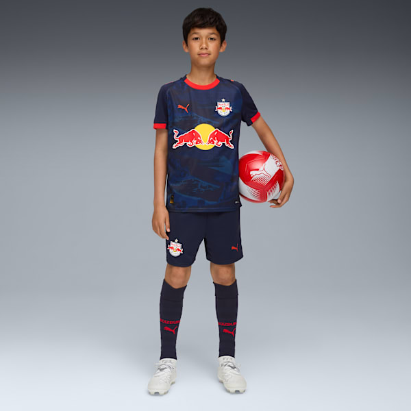 A child poses with a football against a grey backdrop, wearing a sports kit.