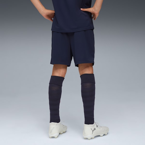 A model stands against grey wearing navy shorts, socks and light-coloured football boots.