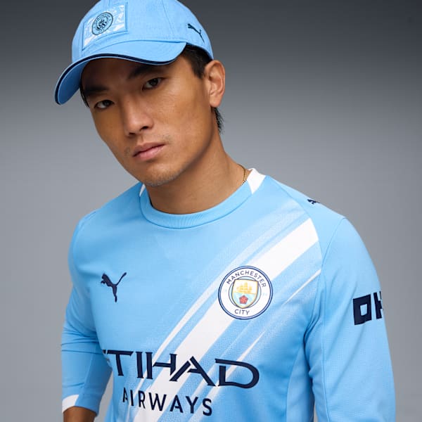 Against grey, a model wears a light blue top, cap, displaying team emblems and sponsor logos.