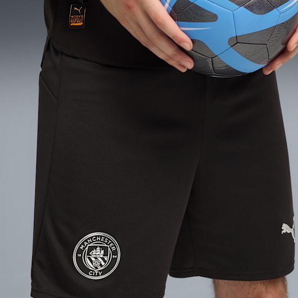 On grey, a person holds a blue football, wearing black shorts with the Manchester City badge.