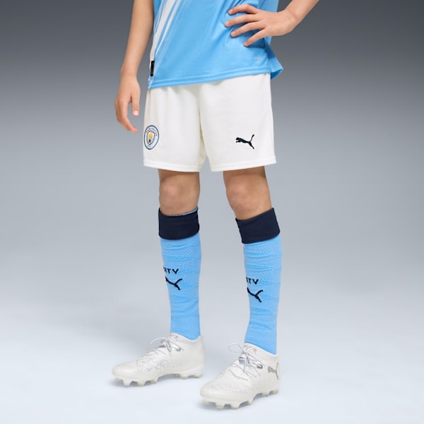 Against a grey backdrop, a model wears a football kit with light blue socks and white shoes.