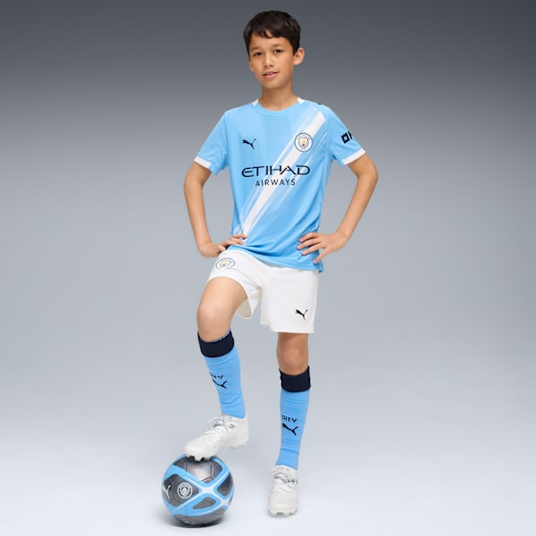 On a grey background, a child poses with a football, wearing a light blue kit, shorts and socks.