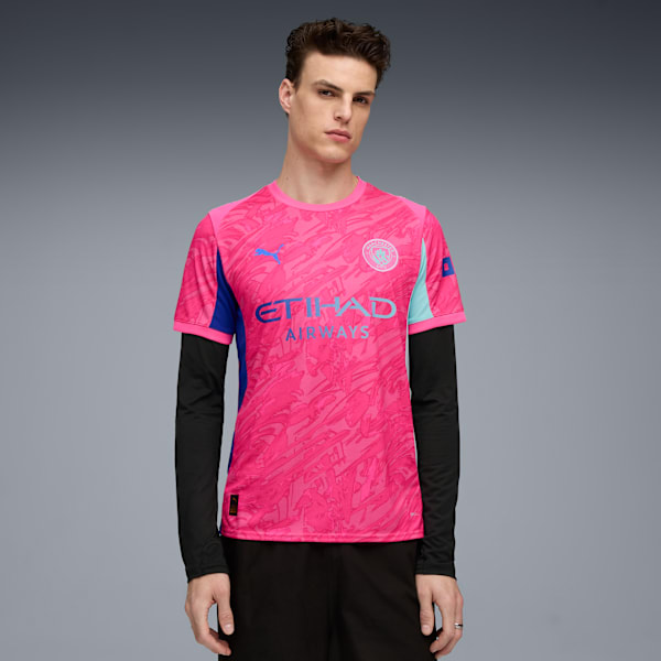 Against grey, a model poses in a pink patterned football shirt with long black sleeves.