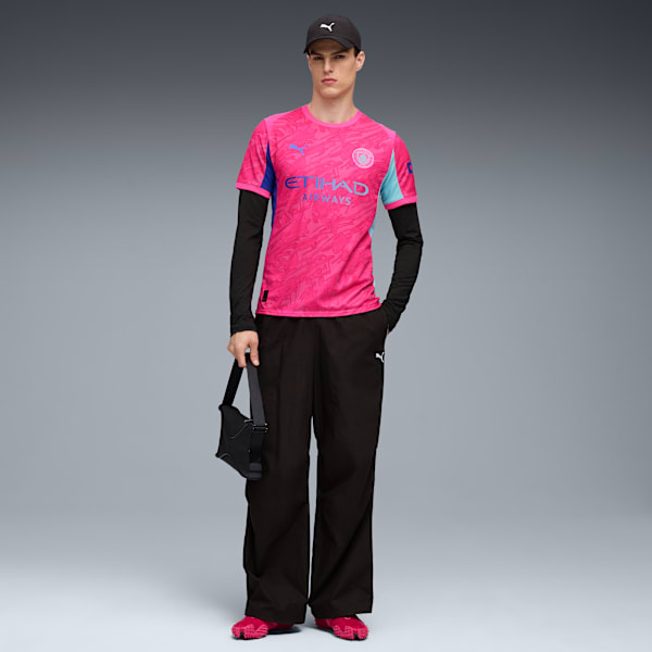 On grey, a model poses in a pink jersey, black trousers, cap, and bright pink shoes.
