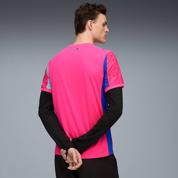 A model with crossed hands, wearing a pink t-shirt with blue details, stands against a grey backdrop.