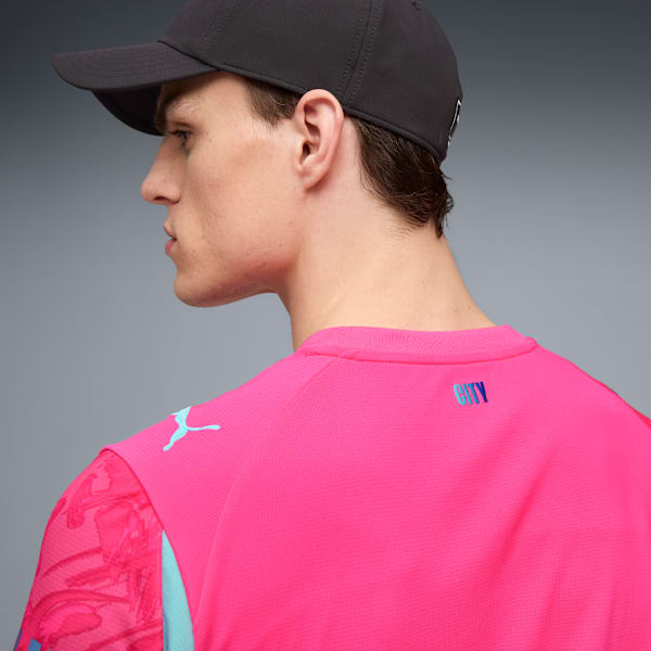 A model faces left against a grey background wearing a pink shirt and a dark cap.