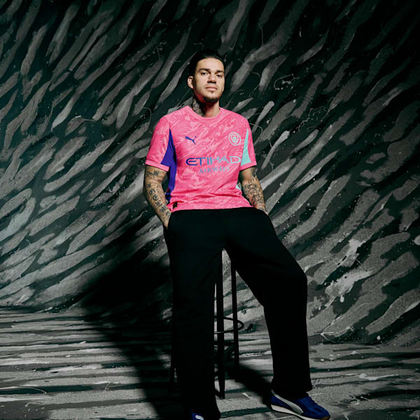 A tattooed model poses against a patterned backdrop, wearing a pink sports shirt and black trousers.