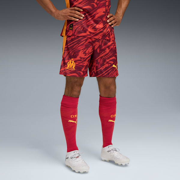 A model poses against grey wearing red patterned shorts, socks and white trainers.