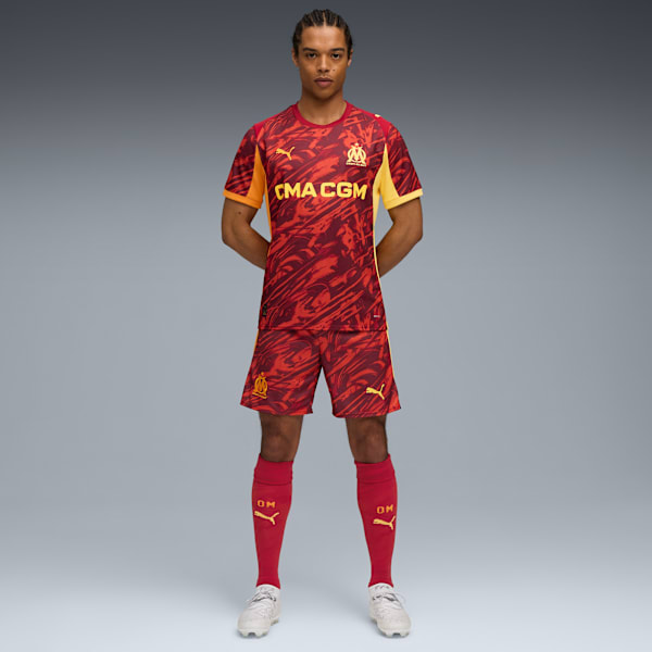 Against grey, a model poses in a red patterned kit with matching socks and trainers.