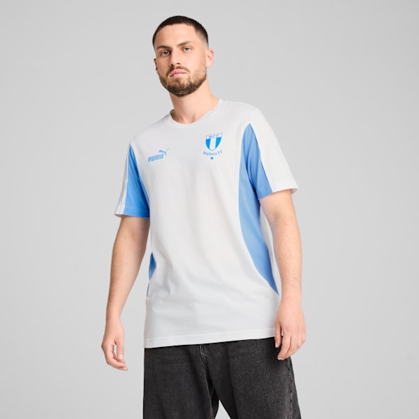 A model poses against a grey backdrop, wearing a white and blue t-shirt with dark trousers.