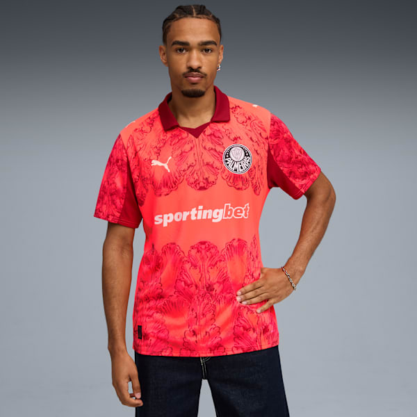 A model poses against grey wearing a coral patterned shirt and dark jeans.