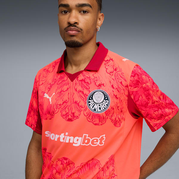 Against grey, a model wears a coral and red patterned football shirt with 'Sportingbet'.