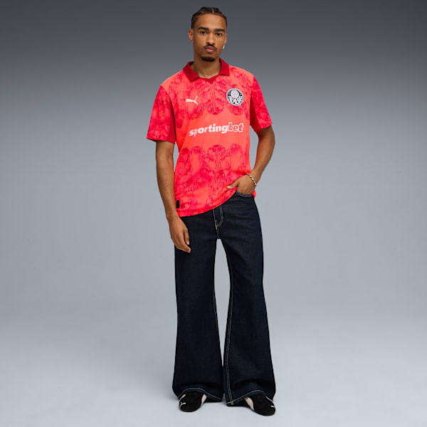 A model stands against grey, wearing a patterned red shirt and wide-leg dark denim jeans.