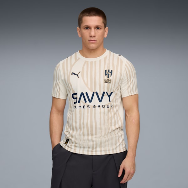 A model stands against grey, wearing a striped t-shirt with logos and dark trousers.