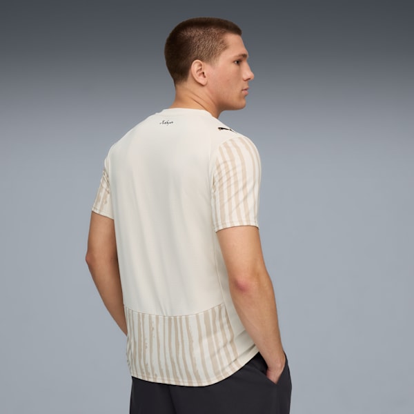 Against grey, a model looks left, wearing a patterned off-white t-shirt and dark trousers.