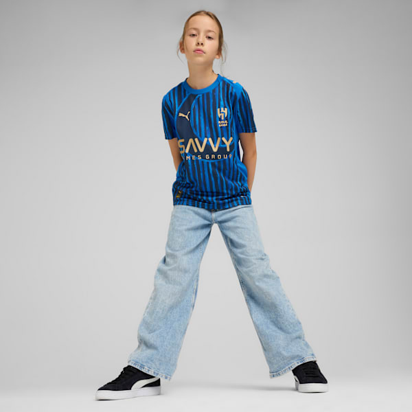 A model stands on a grey background wearing a blue t-shirt, light jeans and black shoes.