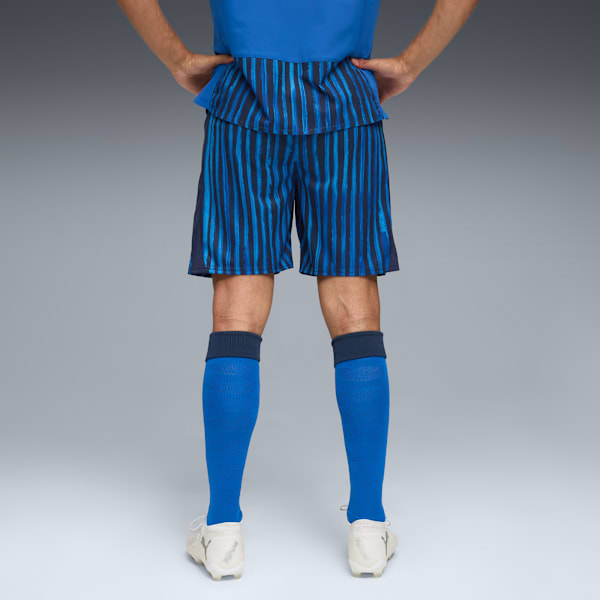 A model stands against grey wearing a blue top, striped shorts, blue socks and white trainers.