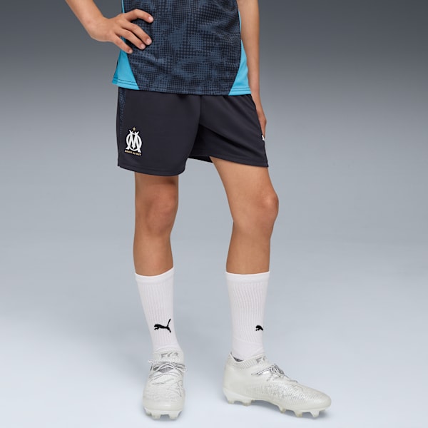 A model poses against grey wearing a shirt, dark shorts, socks and sports shoes.