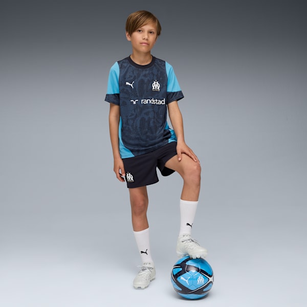 A boy on a grey backdrop, wearing a blue football kit, with his foot on a ball.