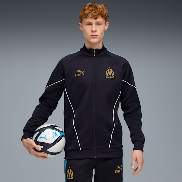 Against grey, a model holds a football wearing a navy tracksuit with light blue and white accents.