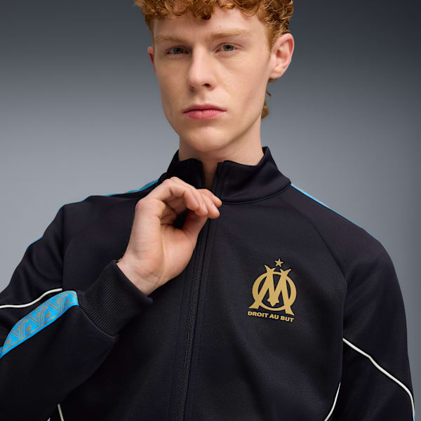 Against grey, a model adjusts a black jacket with gold emblem and blue accents.