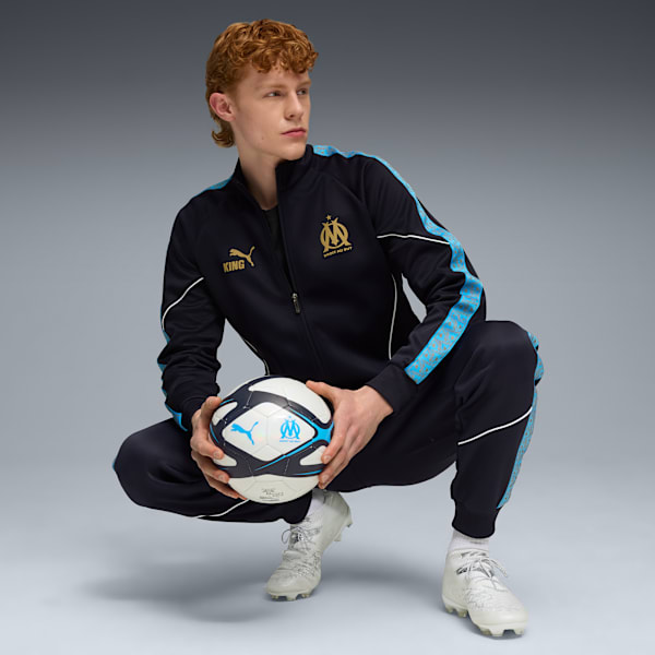 A model crouches on grey, holding a football, wearing a black tracksuit and white football boots.