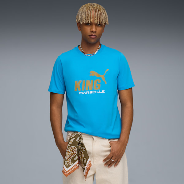 On grey, a model poses in a blue tee with gold lettering and beige trousers.