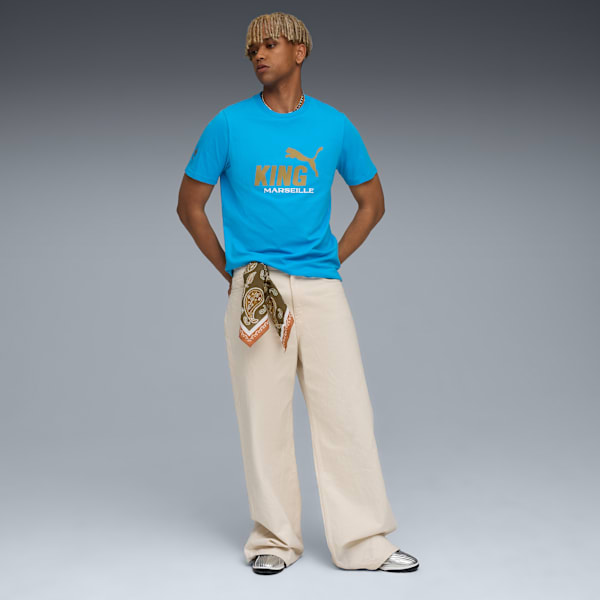 A model poses against grey, wearing a blue 'King Marseille' t-shirt, beige trousers and silver shoes.