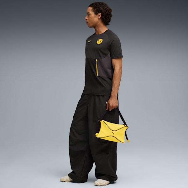 A model stands against a grey backdrop, wearing a dark t-shirt, loose trousers, and holding a yellow bag.