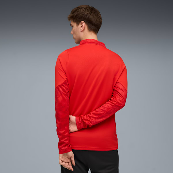 A model faces left against grey, wearing a red top and black trousers with hands behind back.