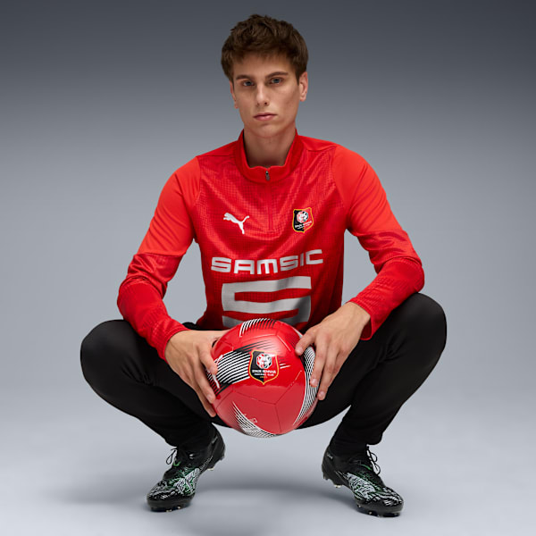 A model squats against grey, holding a red football, wearing a red top and black trousers.
