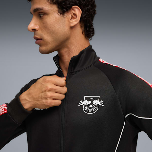 Against grey, a model adjusts a black RB Leipzig track jacket with red stripe detail.