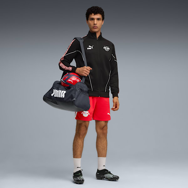 A model with a bag and football stands against a grey backdrop, wearing sportswear.