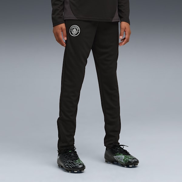 Against grey, a model wears black tracksuit bottoms and football boots.