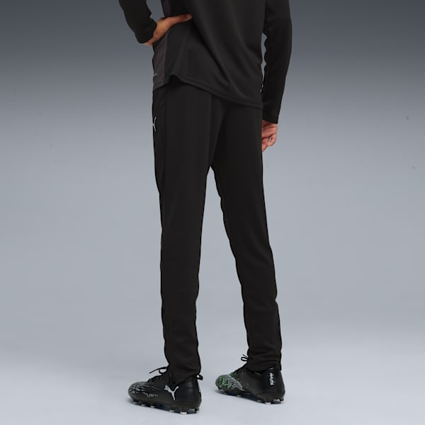 A model on a grey backdrop wears black trousers and football boots, seen from behind.