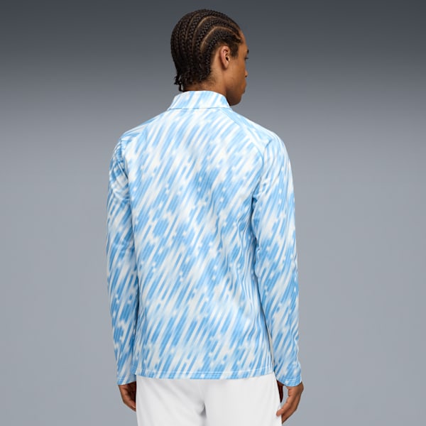 A model poses against grey, wearing a blue patterned long-sleeve shirt and white trousers.