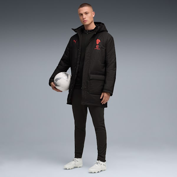Against grey, a model poses holding a football, wearing a black jacket, trousers and white trainers.