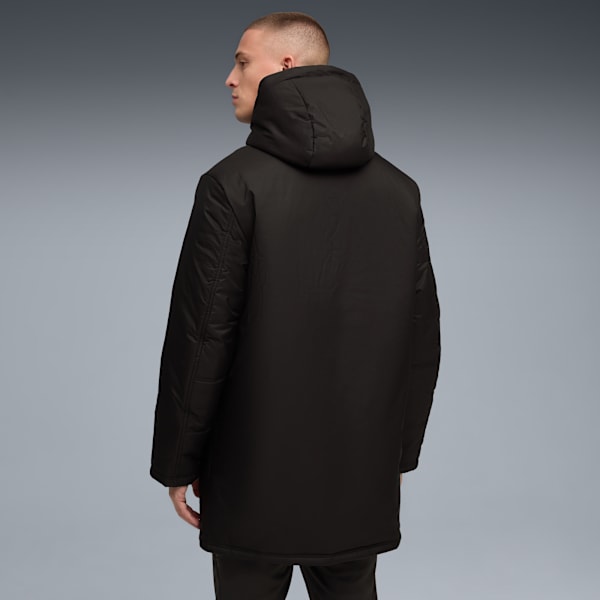 A model faces away on a grey backdrop, wearing a dark hooded coat and trousers.