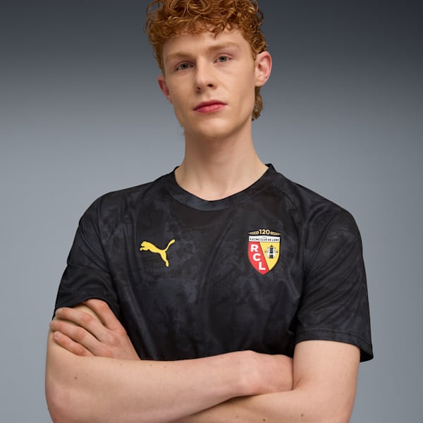 On grey, a model with crossed arms wears a black t-shirt with Puma and club logos.