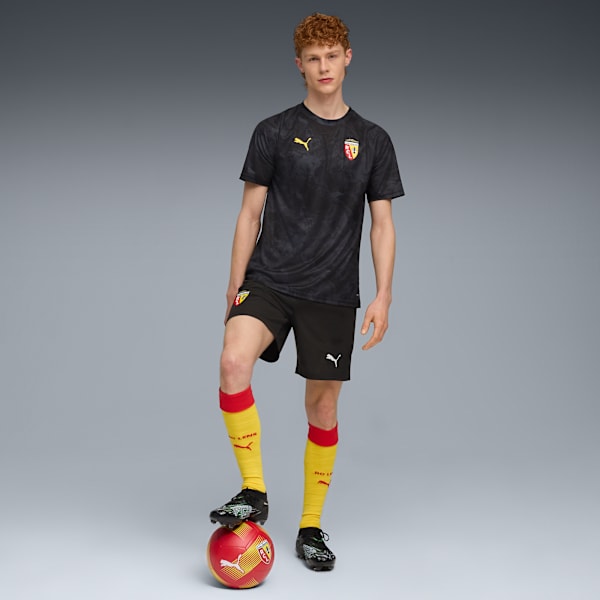 Against a grey backdrop, a model poses with a football, wearing a black kit.