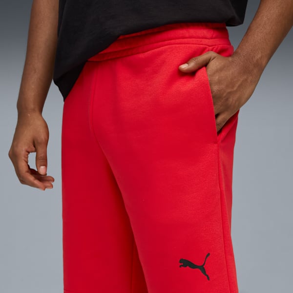 Against grey, a person in red shorts and a black top puts their hand in a pocket.