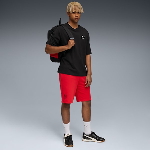 Against grey, a model stands wearing a black tee, red shorts, shoes, and a backpack.